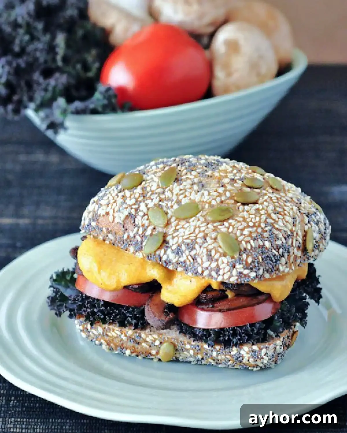 A cheesy mushroom melt sandwich with curly kale, tomato slices, mushrooms and cheese sauce on a seeded bun. Sandwich sits on a light blue plate, with a bowl of tomatoes and mushrooms sitting behind it.