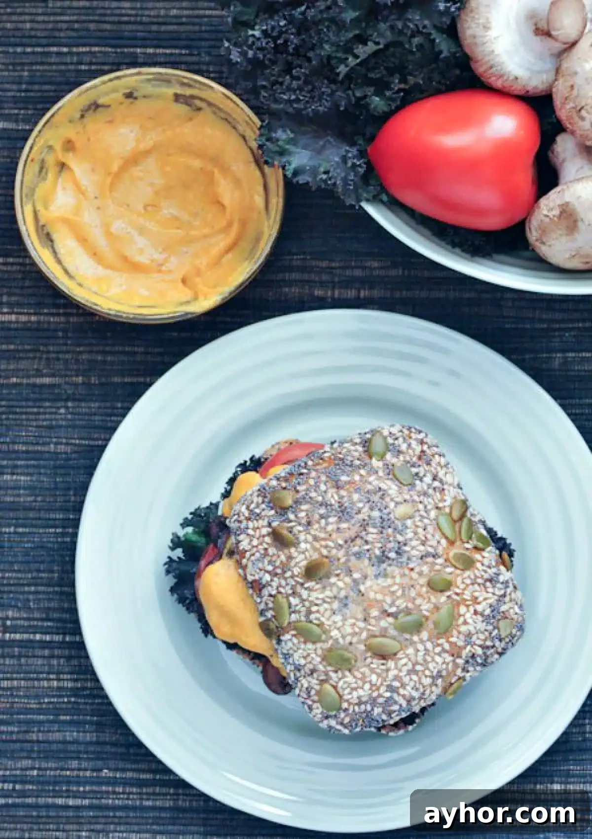 Overhead view of a cheesy mushroom melt sandwich with curly kale, tomato slices, mushrooms and cheese sauce on a seeded bun. Sandwich sits on a light blue plate, with a bowl of tomatoes and mushrooms and a smaller bowl of cheese sauce sitting next to the sandwich.