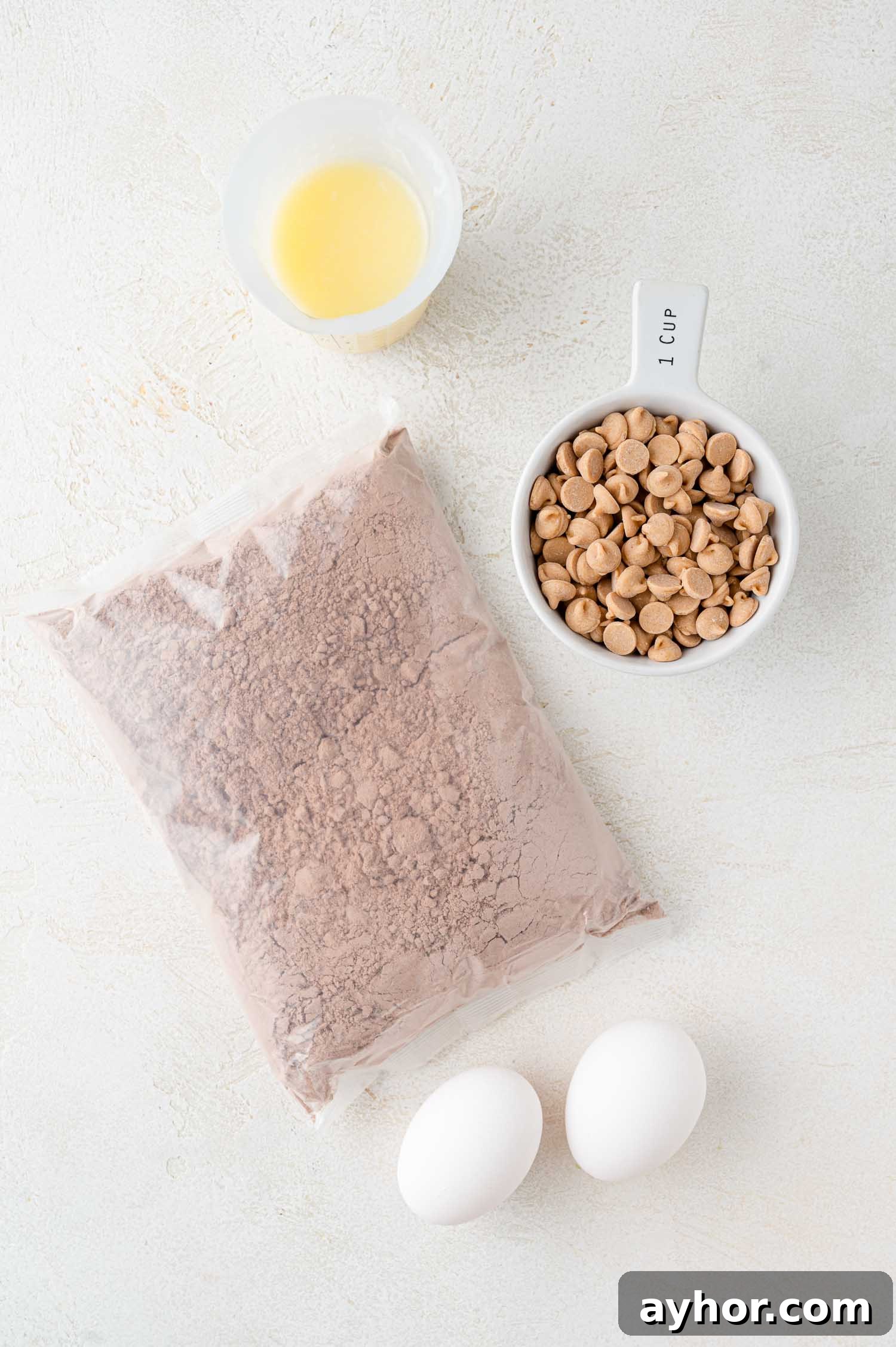 A bag of chocolate cake mix, two eggs, a cup of peanut butter chips, and a small cup of melted butter on a white surface.
