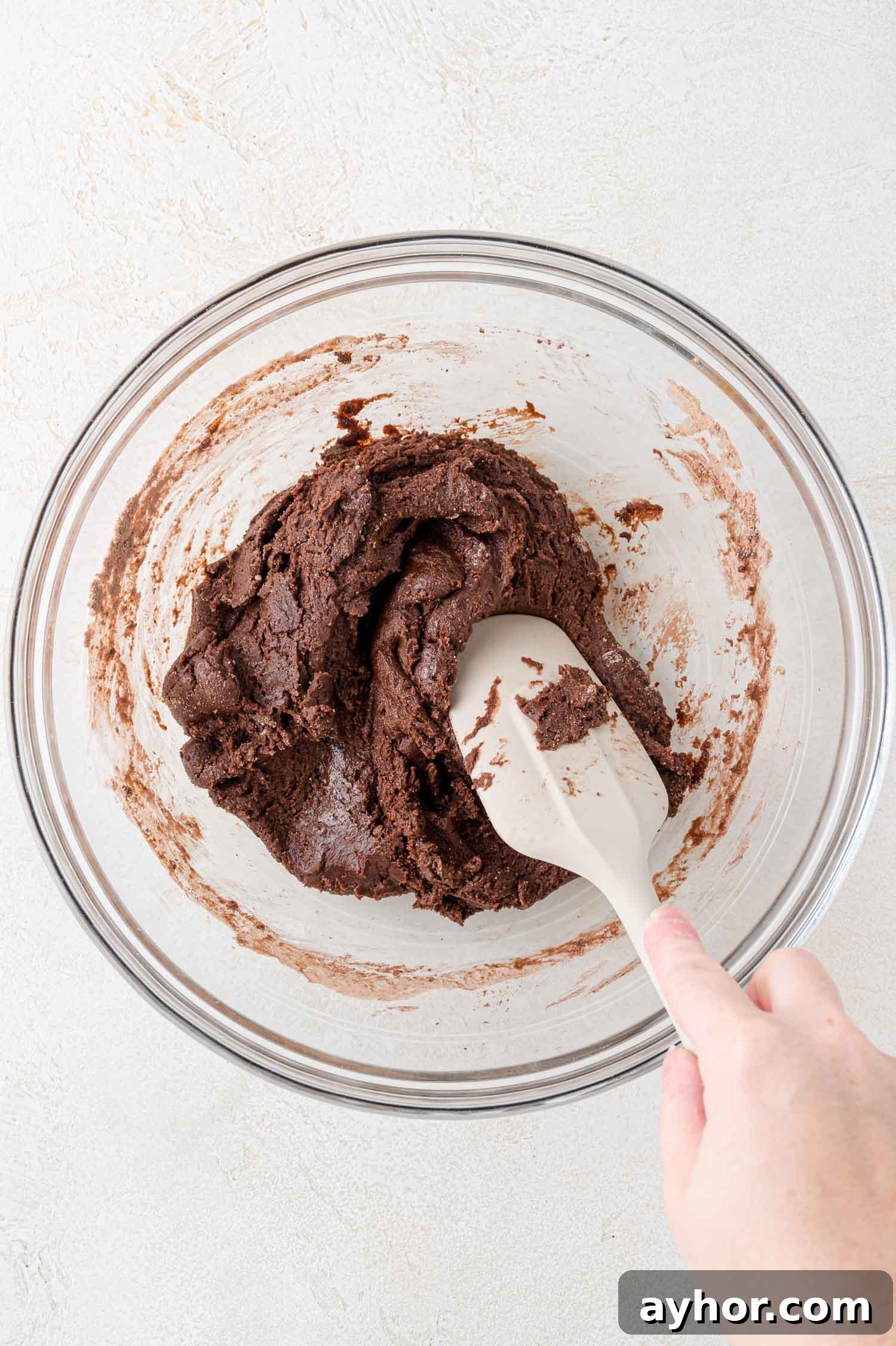 A hand holds a spatula mixing thick chocolate cookie dough in a clear glass bowl on a light-colored surface.