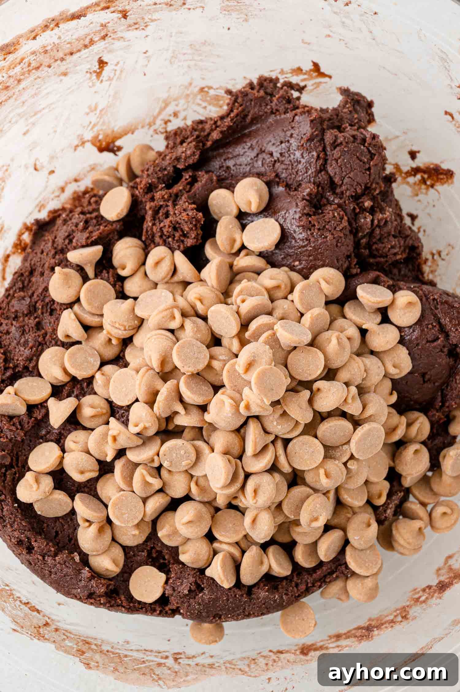 A mixing bowl with chocolate cookie dough topped with a pile of peanut butter chips, ready to be combined.