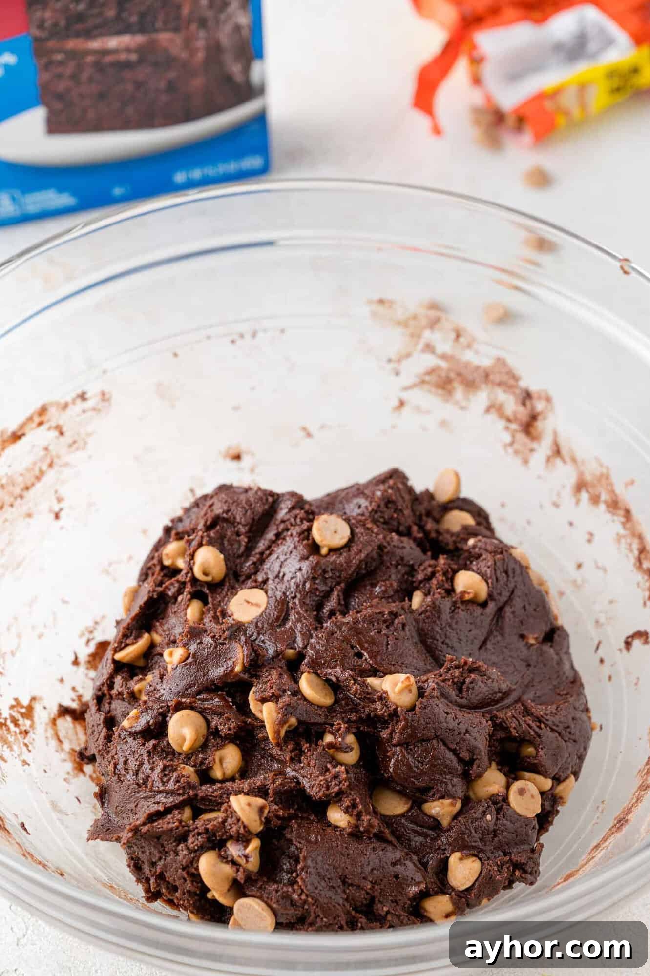 A glass bowl filled with chocolate cookie dough mixed with peanut butter chips sits on a light surface; a box and candy wrapper are in the background.
