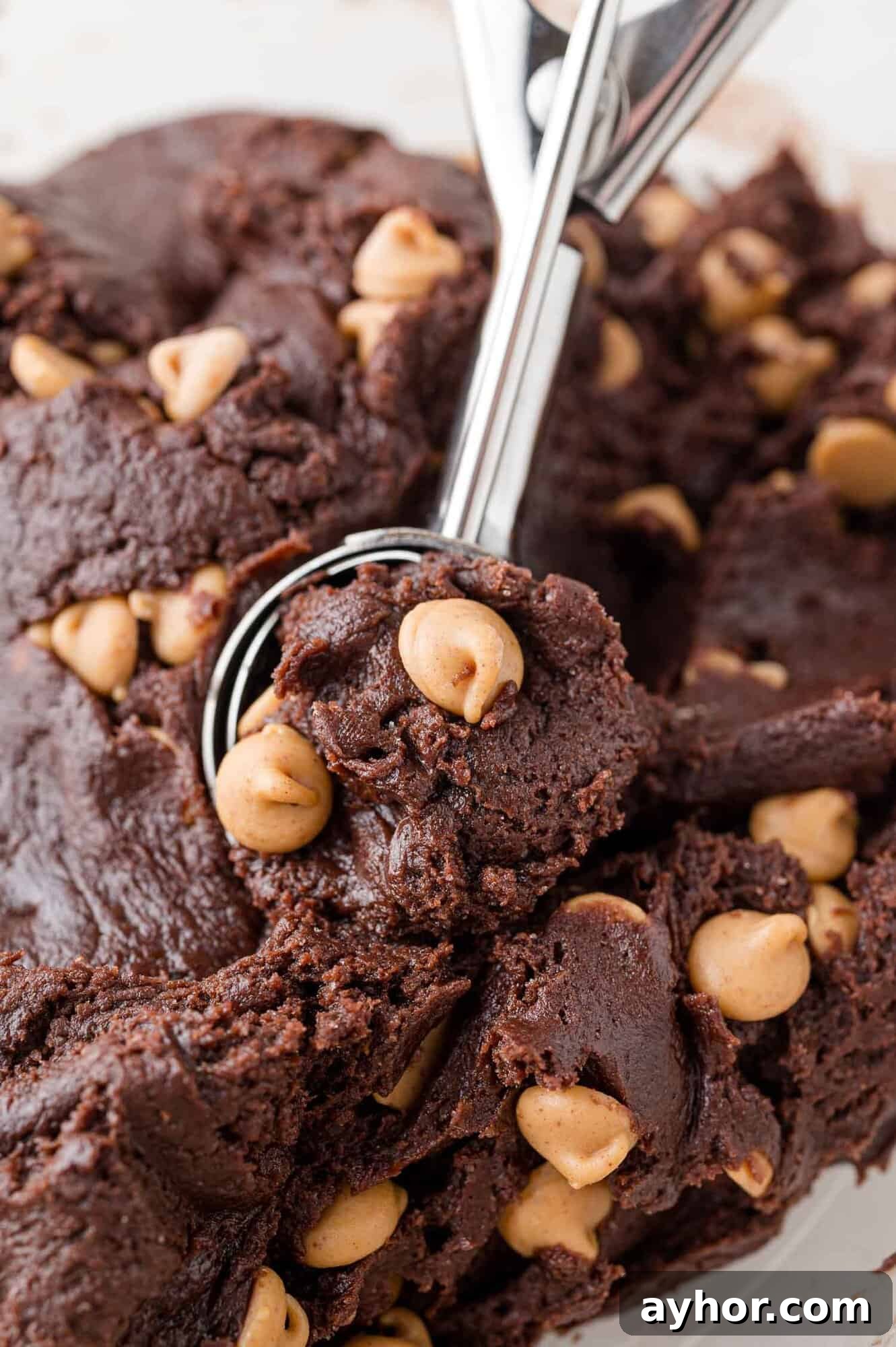 A metal scoop holds a portion of chocolate cookie dough with peanut butter chips, surrounded by more dough in the background.