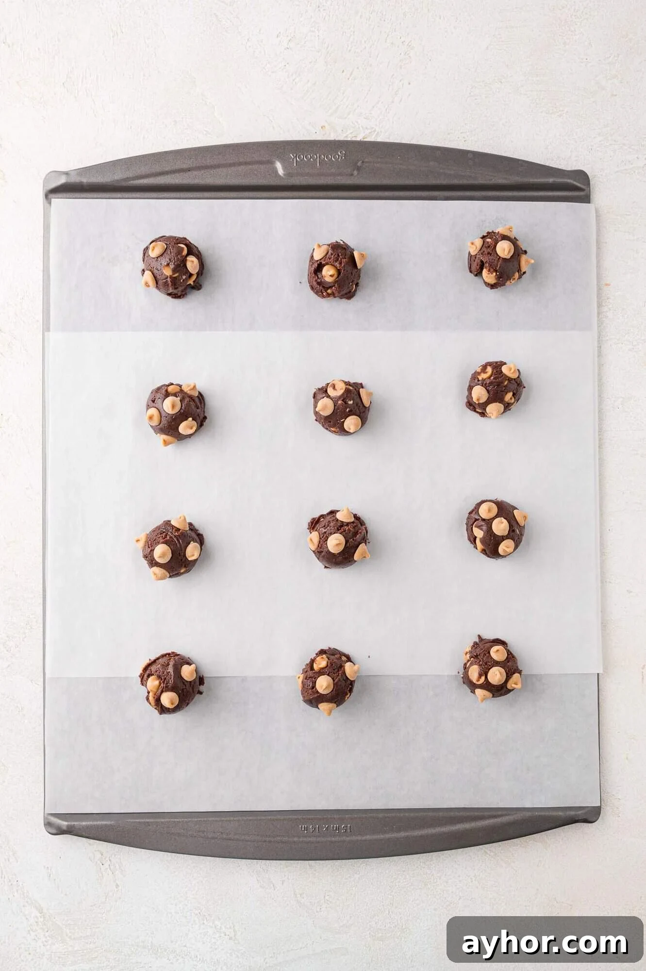 A baking sheet lined with parchment paper holds twelve evenly spaced scoops of chocolate cookie dough with white chocolate chips.
