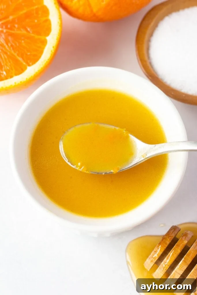 Honey Orange Vinaigrette with 6 Simple Ingredients 2 A white bowl with orange dressing in it, with a spoon dipping a bit of dressing out. It's surrounded by a half an orange, a small wood bowl with kosher salt, and a honey stick dripping with honey.