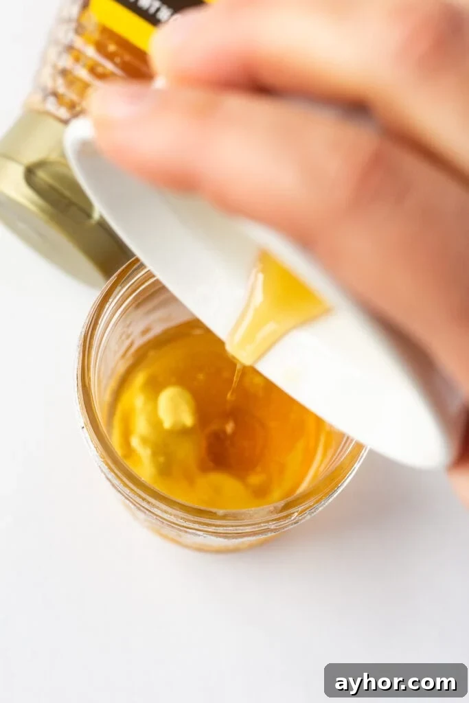 Honey Orange Vinaigrette with 6 Simple Ingredients 6 Pouring honey from a small white bowl into a small mason jar that's has mustard and oil in it.