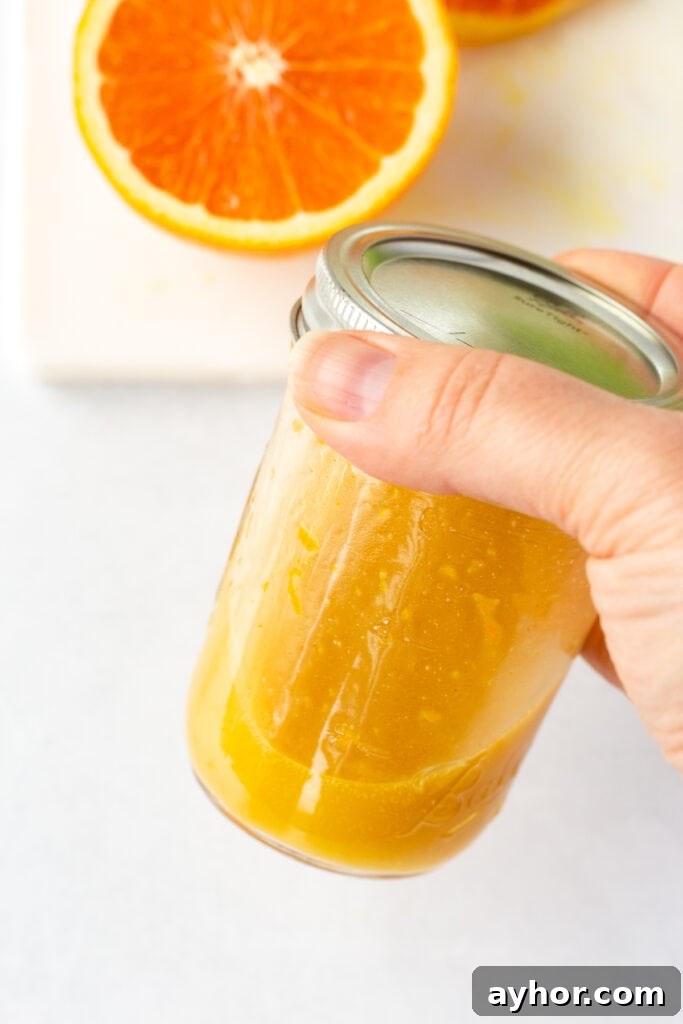 Honey Orange Vinaigrette with 6 Simple Ingredients 8 A hand holds a small mason jar with a lid containing orange dressing. Half an orange on a white cutting board is in the background.