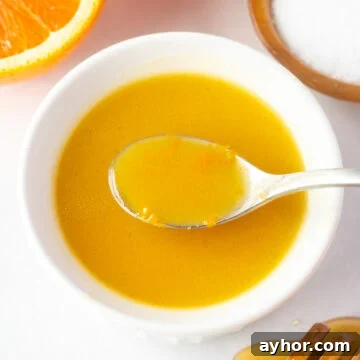 Honey Orange Vinaigrette with 6 Simple Ingredients 10 A white bowl with orange dressing in it, with a spoon dipping a bit of dressing out. It's surrounded by a half an orange, a small wood bowl with kosher salt, and a honey stick dripping with honey.