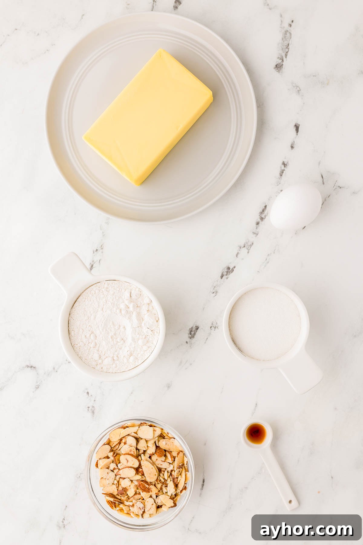 Sand Dollar Cookie Recipe: Crisp Shortbread Cookies Shaped Like Sand Dollars 5 Butter on a plate, an egg, flour, sugar, chopped nuts in a bowl, and a spoon of vanilla extract arranged on a marble countertop.