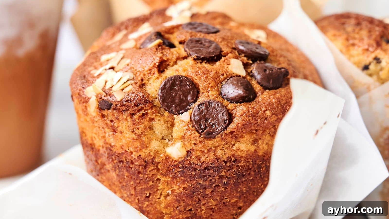 29 Breakfast Recipes to Make Your Mornings Feel Romantic 26 Close-up of a freshly baked muffin topped with chocolate chips and oats, wrapped in a paper liner. A blurred drink and other baked goods are visible in the background.