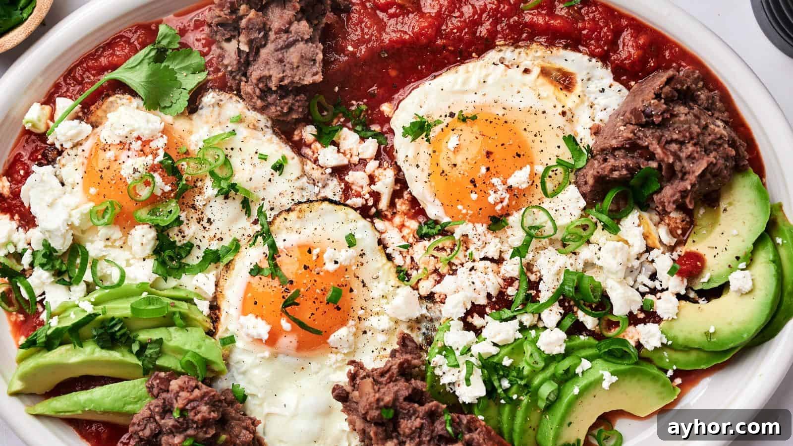 29 Breakfast Recipes to Make Your Mornings Feel Romantic 29 A plate of huevos rancheros with sunny-side-up eggs, avocado slices, refried beans, tomato sauce, crumbled cheese, and garnished with green onions and cilantro.