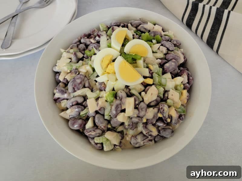 Kidney bean salad garnished with hard boiled egg slices in a white bowl next to plates with two forks and a cloth napkin