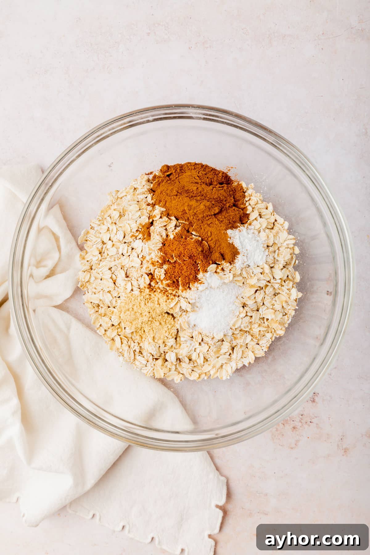 A glass mixing bowl with gluten-free oats topped with cinnamon, nutmeg, ginger, salt, and baking powder before whisking together.