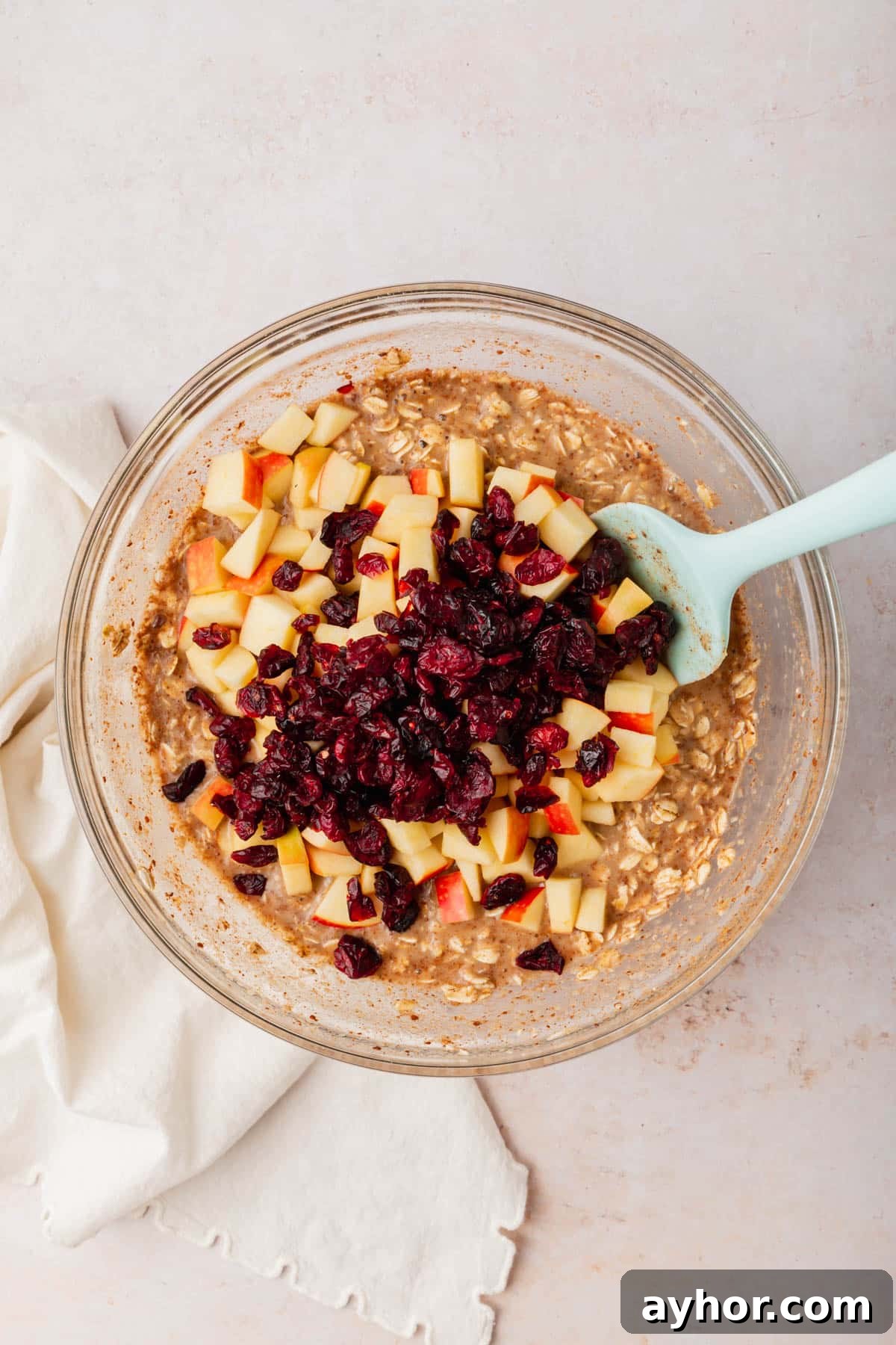 A glass mixing bowl with an oatmeal mixture topped with diced apples and dried cranberries before mixing together.