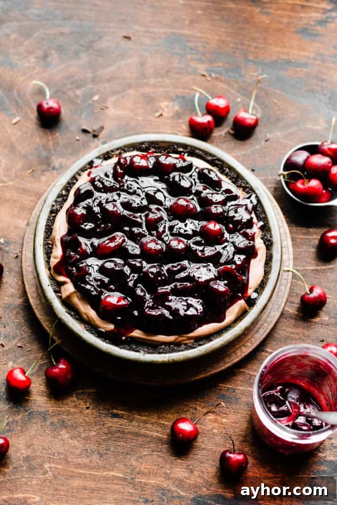 No-Bake Black Forest Chocolate Cherry Pie Recipe 6 Chocolate mousse filling in a chocolate pie crust, topped with cherry sauce, on a wooden surface dotted with fresh cherries