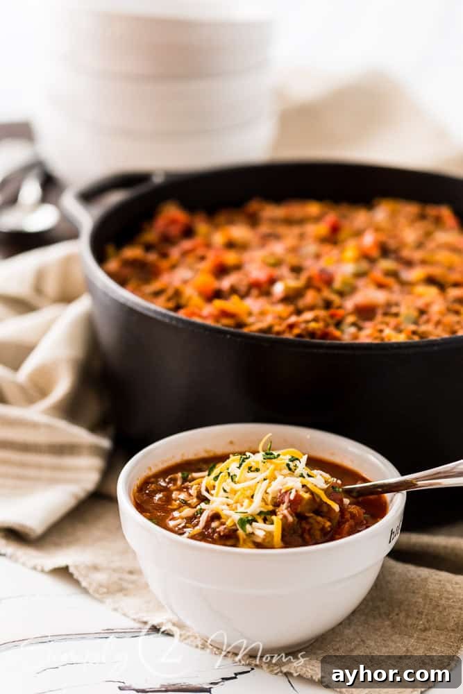 All-Meat Keto Chili Recipe (THM-S Friendly) 3 Bowl of keto chili garnished with shredded cheese in front of a cast iron dutch oven filled with an all meat chili without beans on a table with bowls and spoons