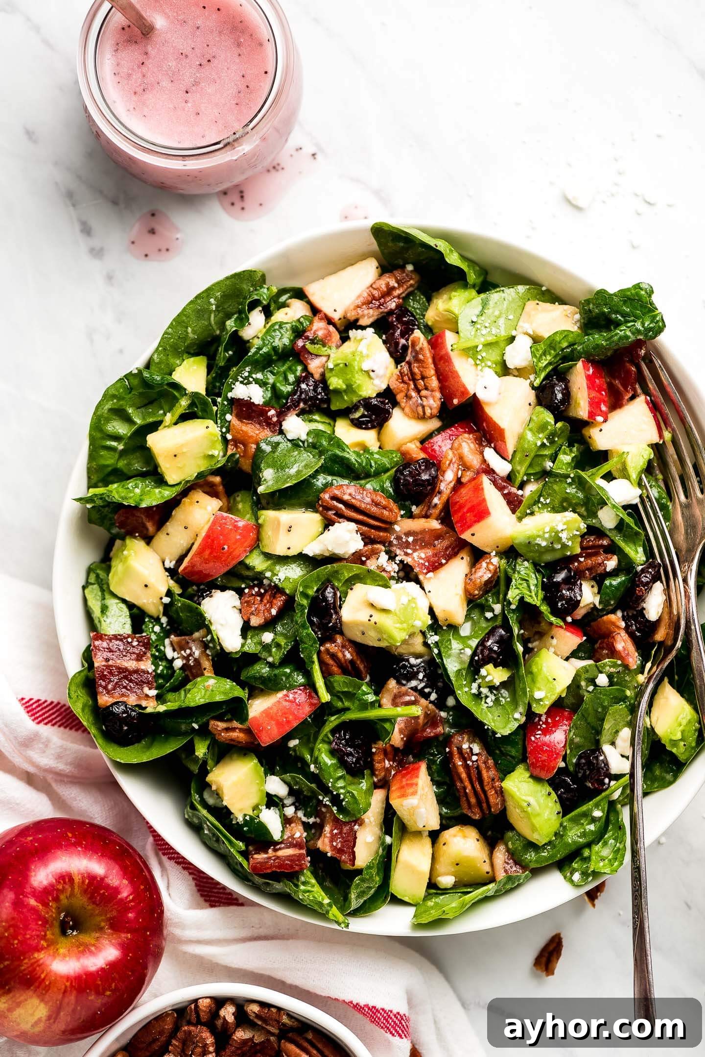 Apple & Bacon Spinach Salad tossed in a poppy seed dressing in a large bowl with an apple and a jar of pink poppy seed dressing to the side.