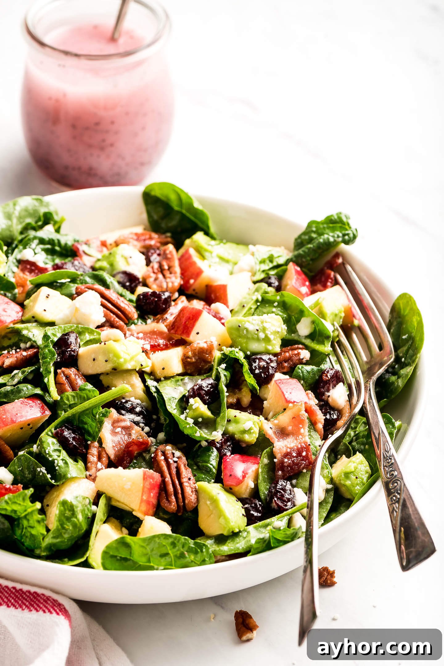 A tossed poppy seed spinach salad with two fork in the bowl with a jar of poppy seed dressing in the background.