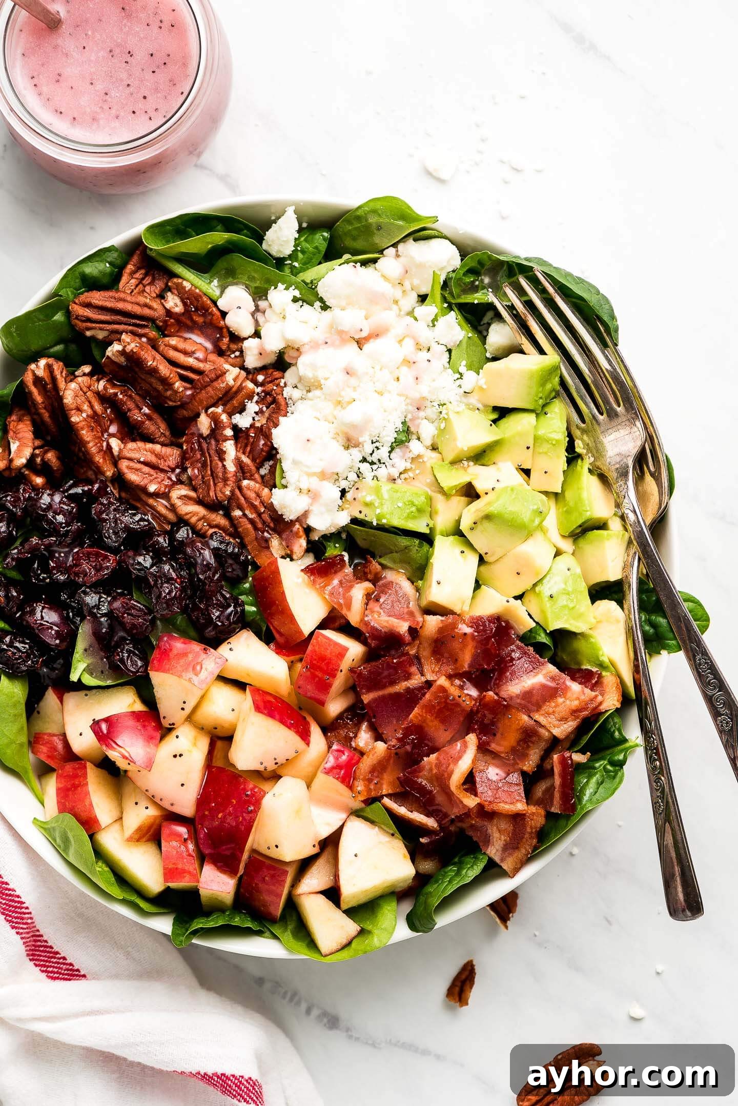 A bowl of baby spinach topped with pecans, feta cheese, diced avocado, chopped bacon, diced apple, and dried cranberries.