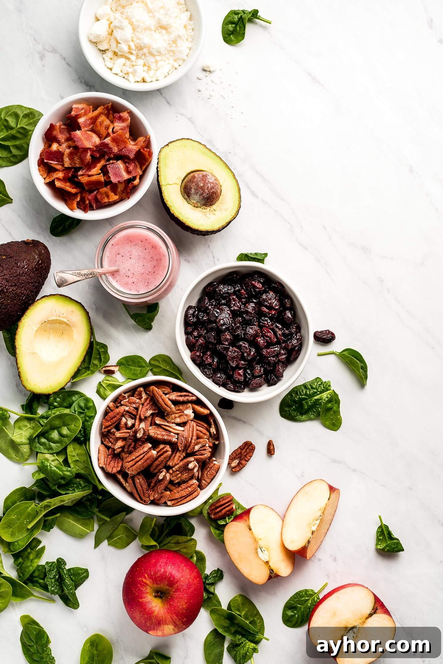 A marble surface topped with various ingredients- feta cheese, bacon, dried cranberries, toasted pecans, poppy seed dressing, apples, and avocado.