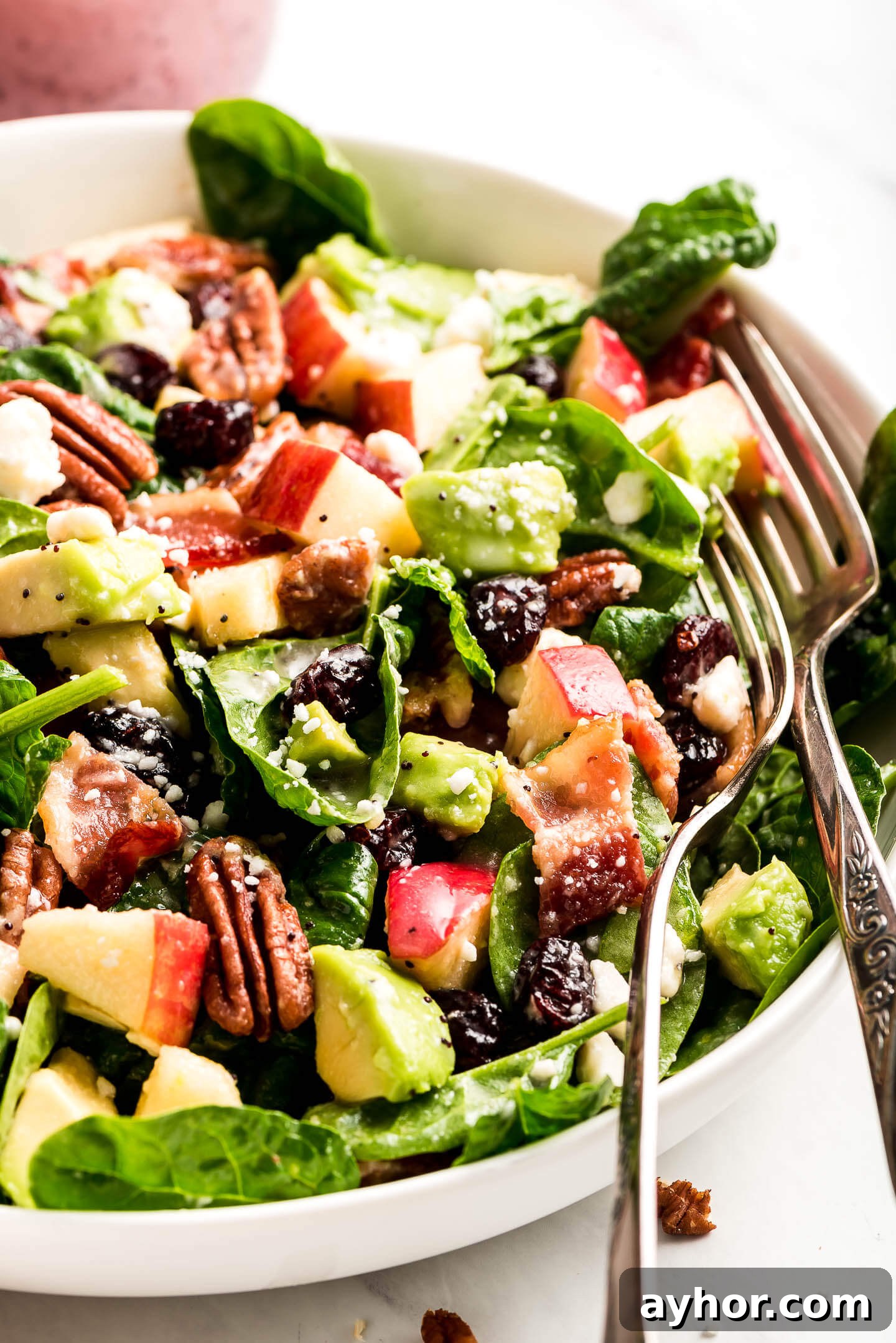 A close-up shot of Apple & Bacon Spinach Salad tossed in poppy seed dressing with two forks in the side of the bowl.