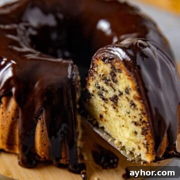 Baked chocolate chip pound cake with chocolate ganache on round wooden cake platter. One slice being removed with a spatula.