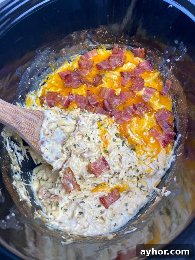 Slow Cooker Chicken with Bacon and Ranch Flavor 4 Shredded chicken bacon ranch in a crockpot with cheddar cheese and a wooden spoon stirring it together.