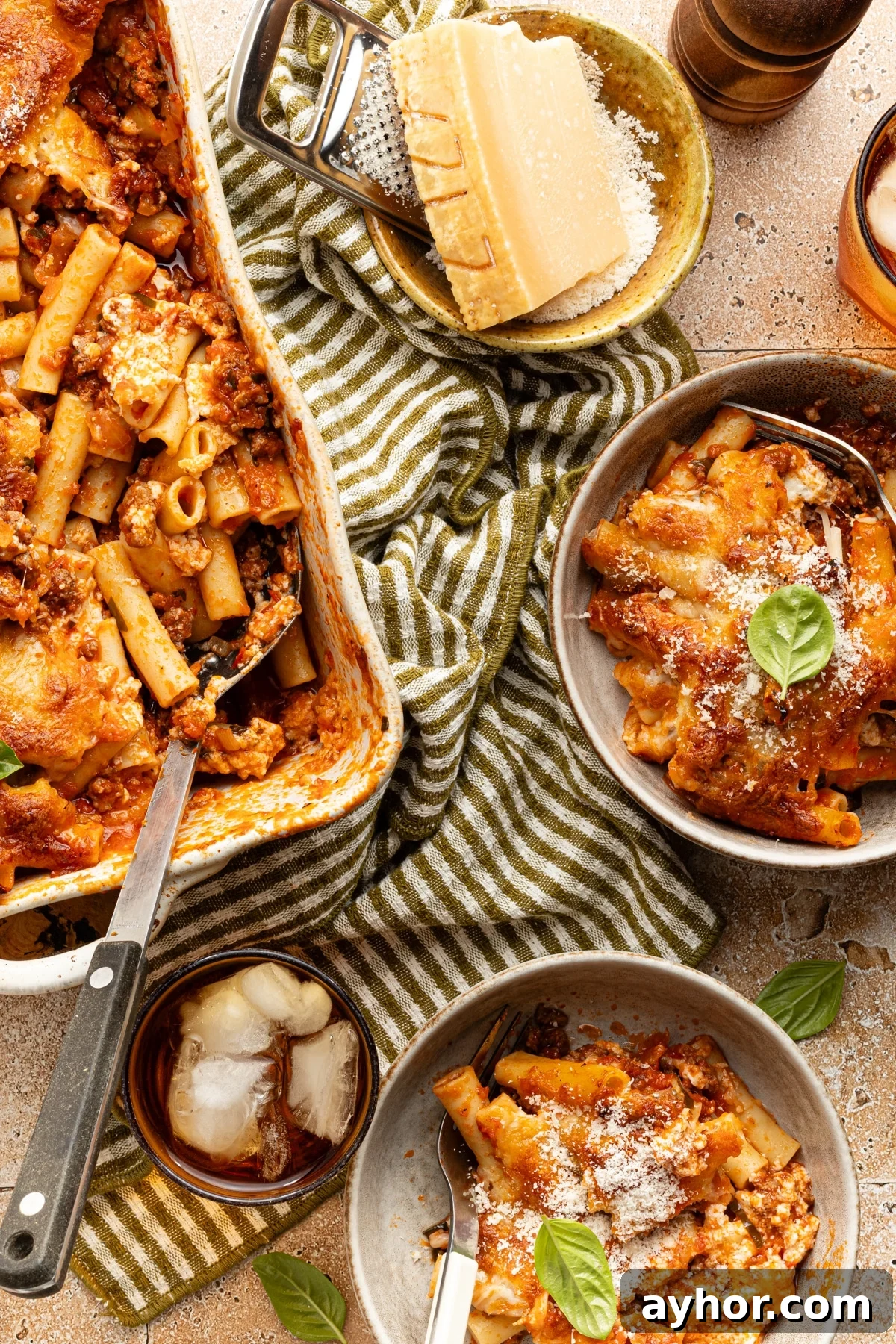 High-Protein Baked Ziti Recipe for Muscle & Meal Prep 11 High protein baked ziti in a large baking dish next to two portioned bowls topped with grated cheese and fresh basil.