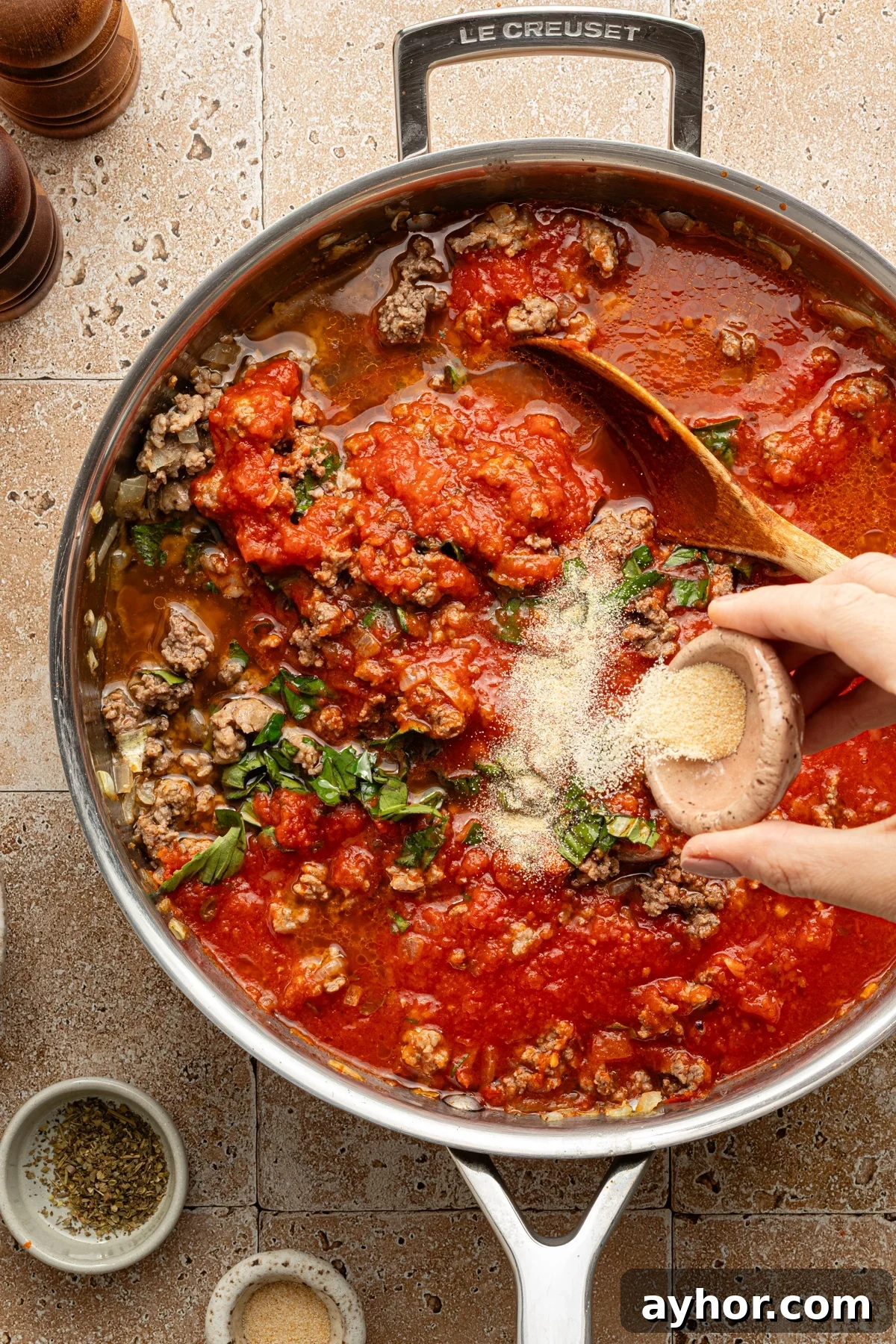 High-Protein Baked Ziti Recipe for Muscle & Meal Prep 5 Seasonings being added by hand to a large skillet with cooked meat, water and tomato sauce.