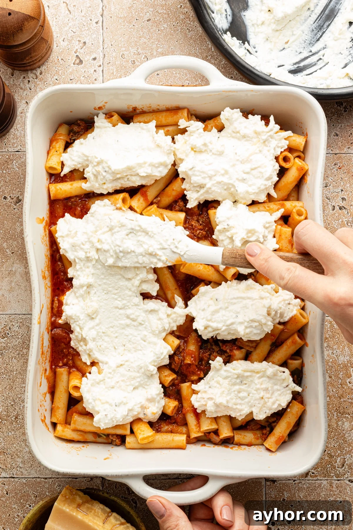 High-Protein Baked Ziti Recipe for Muscle & Meal Prep 8 Cheese mixture dollops being spread over a layer of ziti and meat sauce in a large white baking dish.