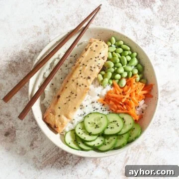 Miso butter salmon in a bowl with rice, edamame, carrots, and cucumber, topped with black sesame seeds, chopsticks rest on the left side of the bowl.