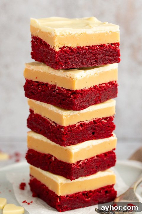 A stack of 5 red velvet millionaire brownies set on two stacked plates with a fork on the side. There are white chocolate hearts, red velvet brownie crumbs and a light brown napkin in the background.