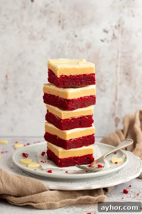 A stack of 5 red velvet millionaire brownies set on two stacked plates with a fork on the side. There are white chocolate hearts, red velvet brownie crumbs and a light brown napkin in the background.