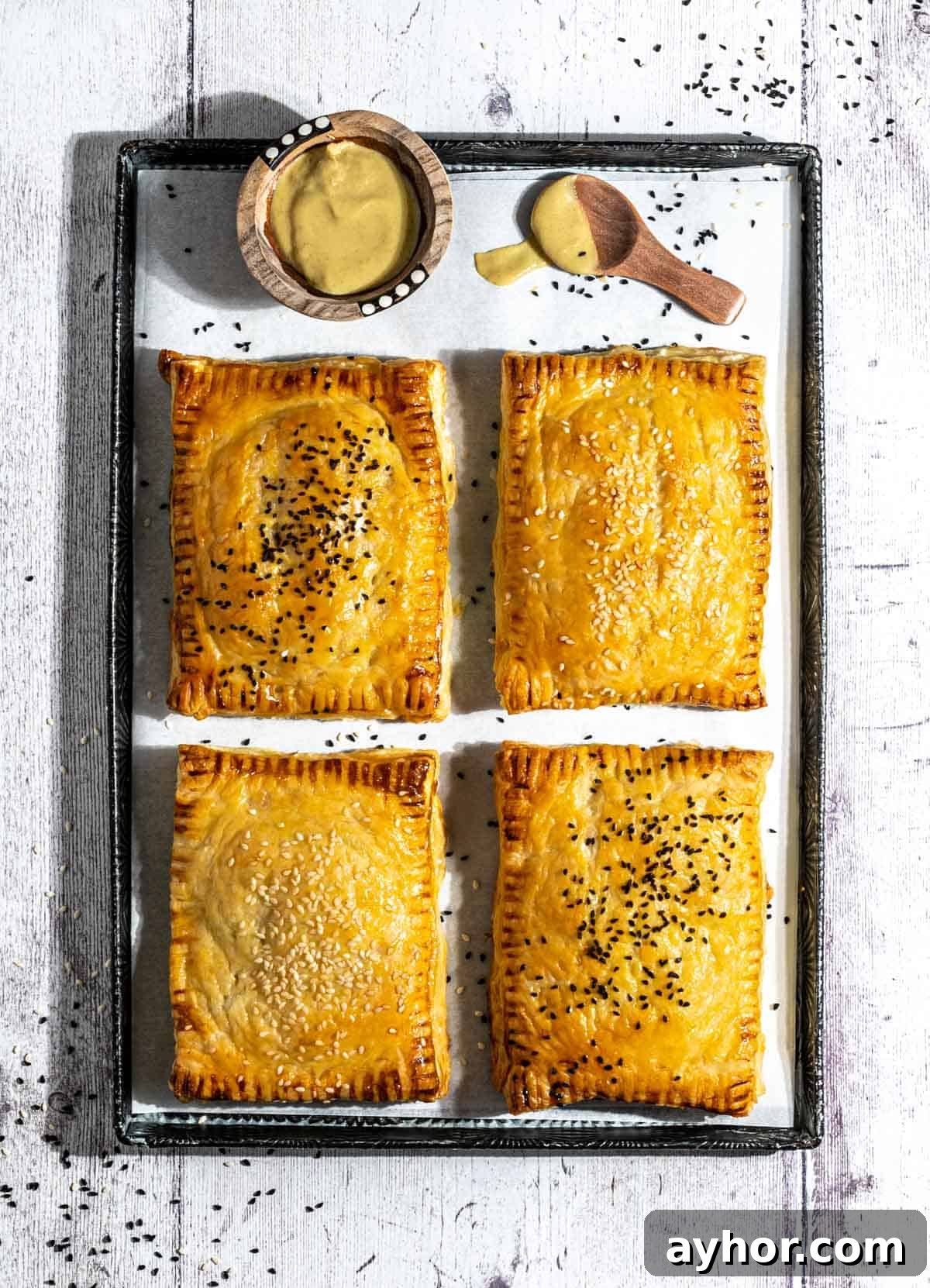 Four cheese and onion pasties on a baking sheet with a small bowl of mustard.
