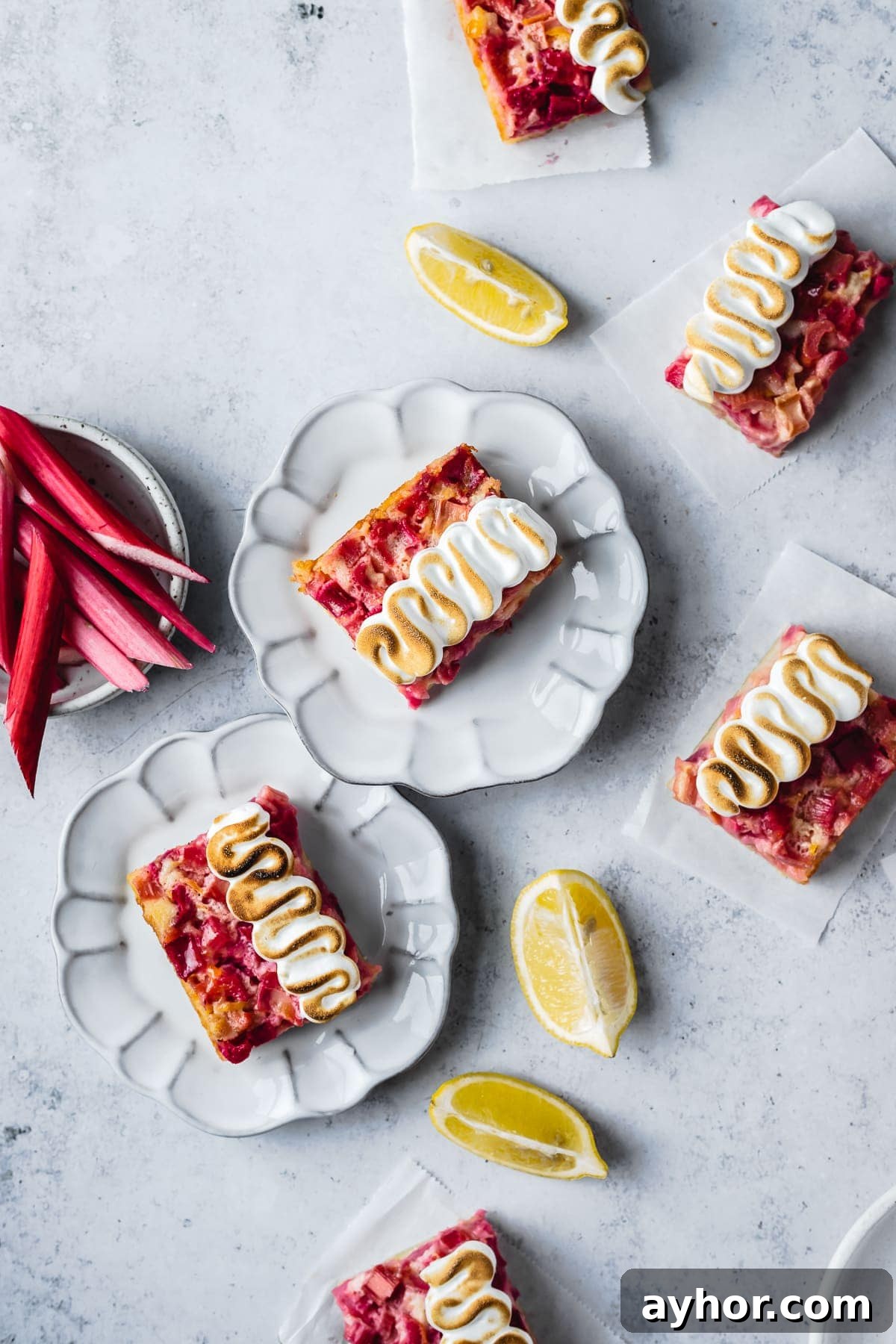 Lemon Rhubarb Bars Recipe with Buttery Shortbread Crust 4 Bar cookies with decorative meringue piping on plates and parchment squares on a white concrete surface