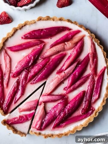Strawberry rhubarb panna cotta tart with a slice cut into it on a grey marble background with strawberries, rhubarb and plates nearby