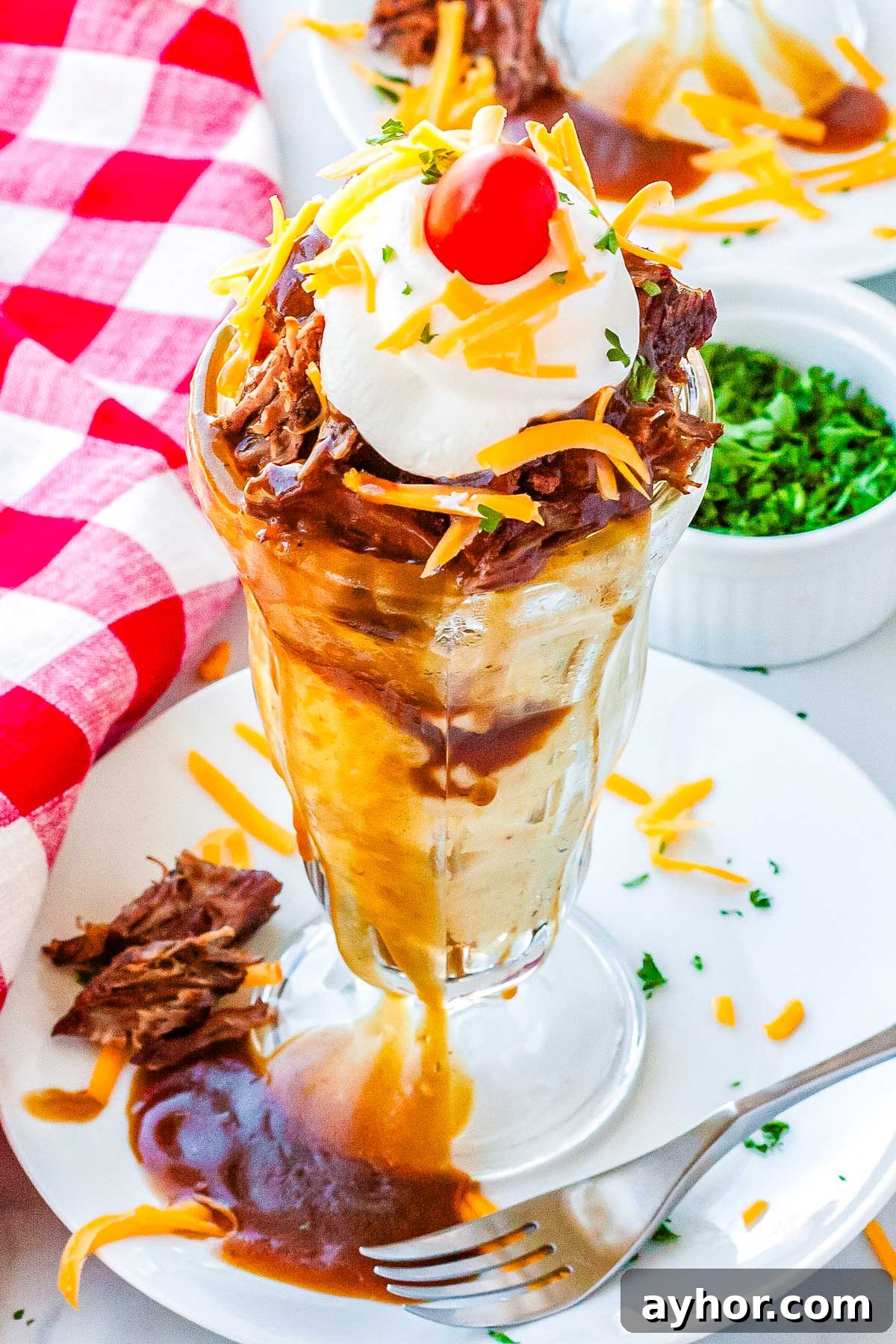 A hot beef sundae in a clear sundae dish topped with shredded cheese, sour cream, and a cherry tomato.