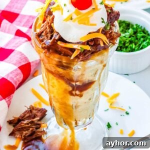 A hot beef sundae in a clear sundae dish topped with shredded cheese, sour cream, and a cherry tomato.
