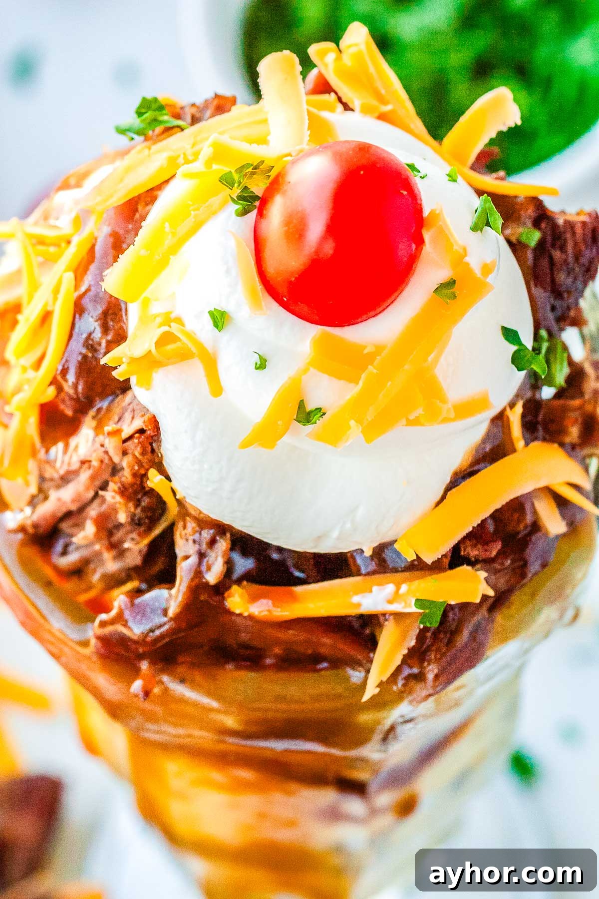 A hot beef sundae topped with shredded cheddar cheese, a dollop of sour cream, a cherry tomato, and parsley.