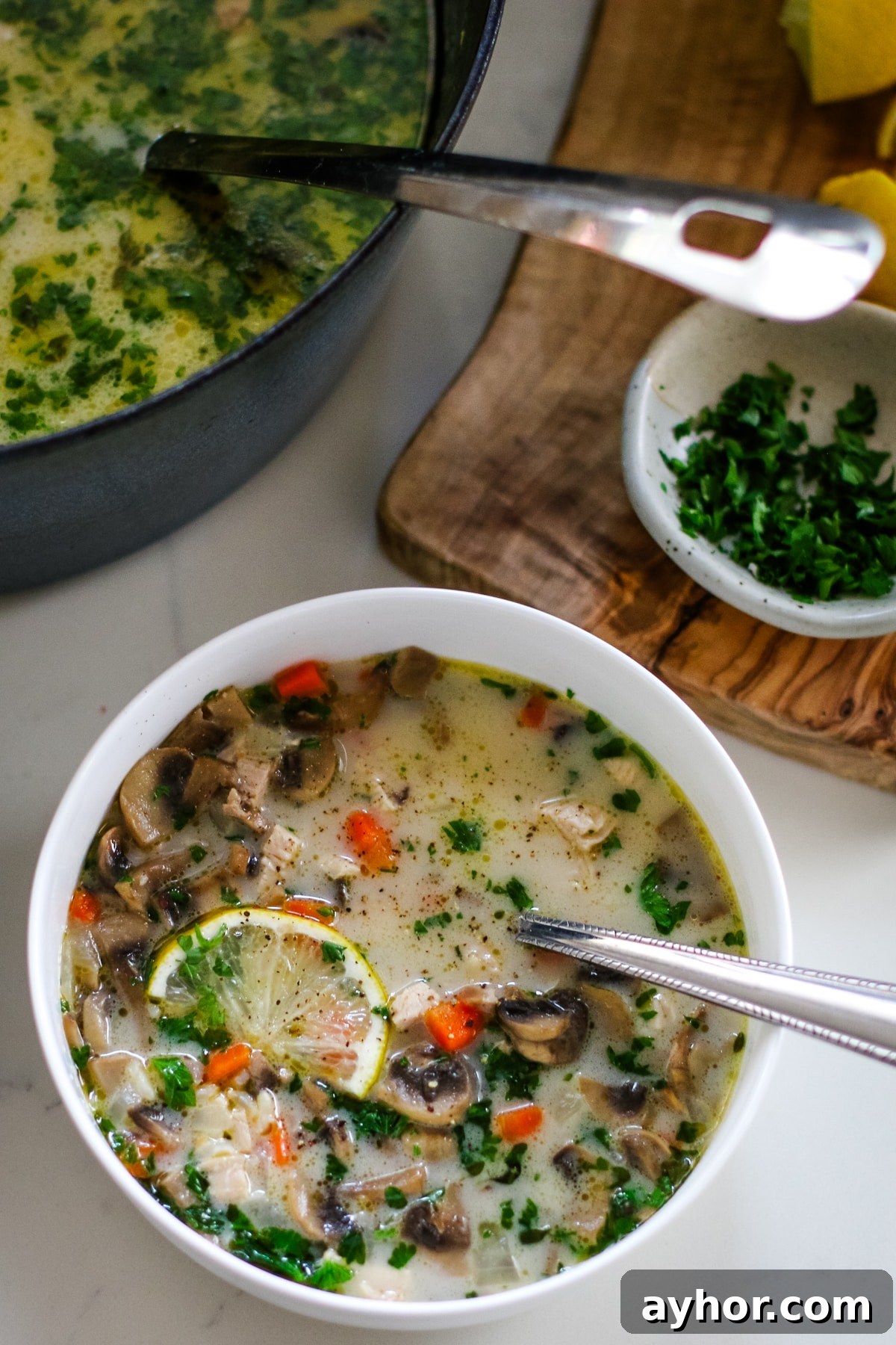 Mushroom, Rice & Turkey Soup from Leftover Roast 13 Turkey soup in a white bowl with a spoon and a slice of lemon, next to a cutting board and a pot full of turkey soup.