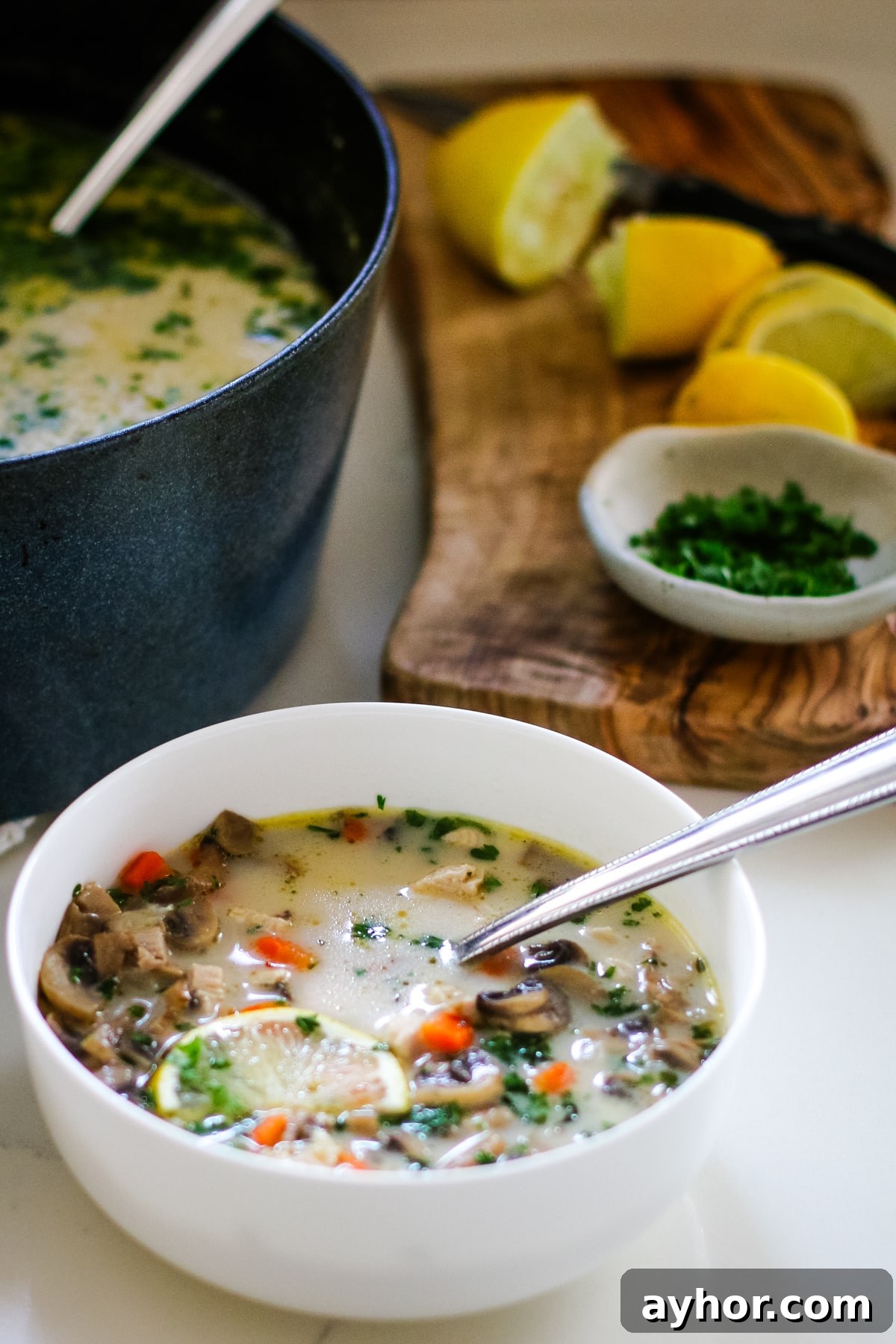 Mushroom, Rice & Turkey Soup from Leftover Roast 6 Turkey soup in a white bowl with a spoon and a slice of lemon, next to a cutting board and a pot full of turkey soup.