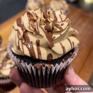 Espresso Chocolate Peanut Butter Cupcakes Recipe 4 A hand holds a chocolate cupcake topped with swirls of light brown frosting and drizzled with chocolate. The background is blurred, showing more cupcakes on a wooden surface.