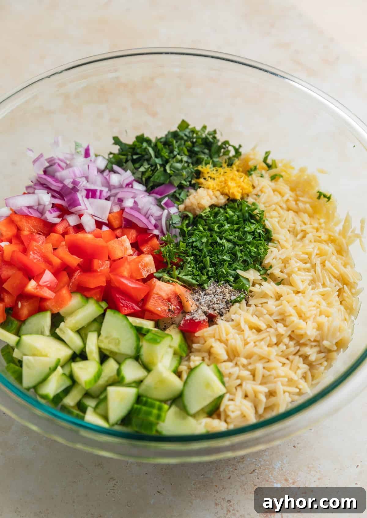 Orzo, lemon zest, cucumber, peppers and other ingredients in glass mixing bowl.