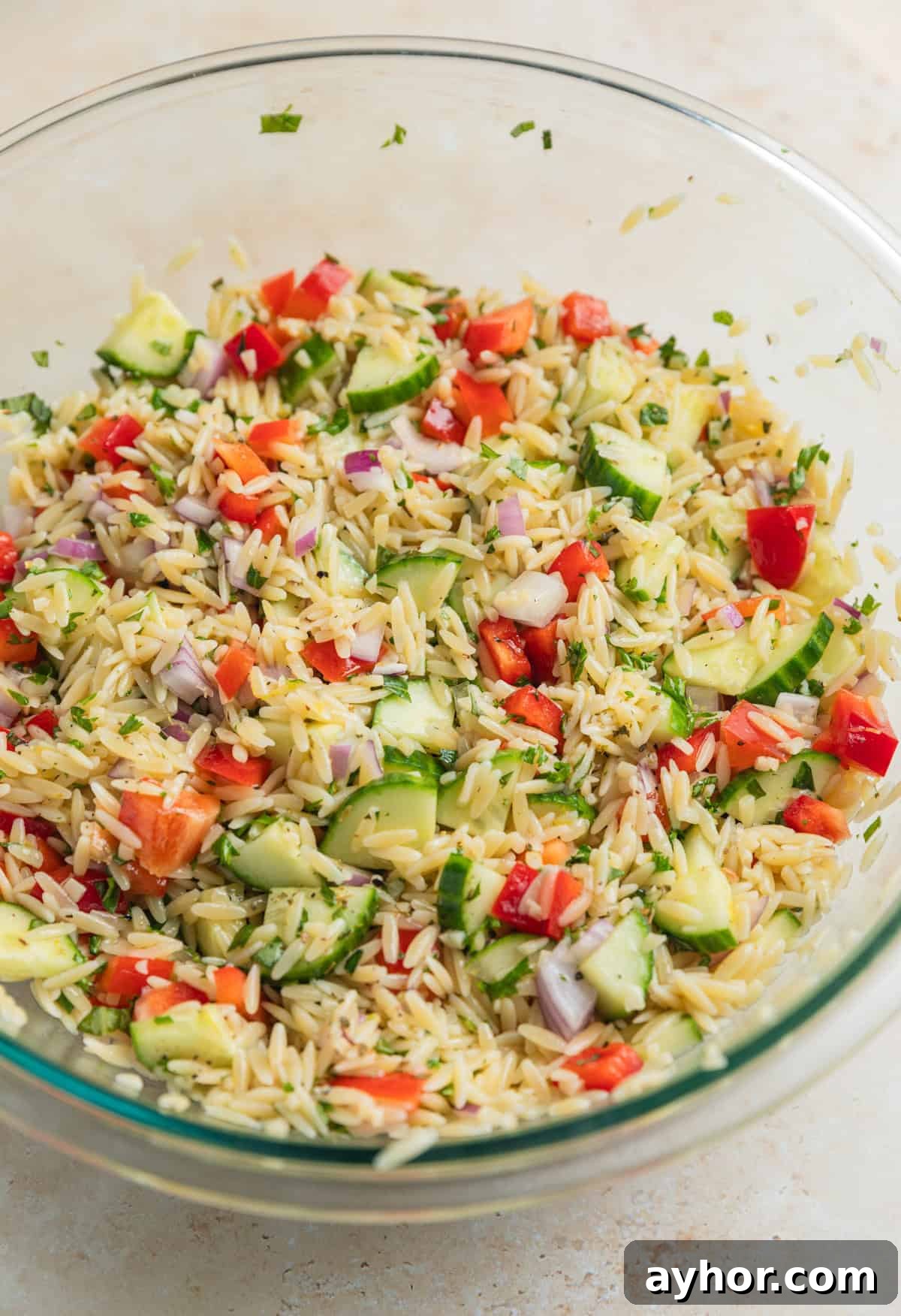 Orzo salad ingredients tossed together in glass mixing bowl before adding feta.