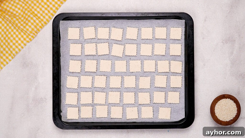 A parchment-lined baking sheet filled with rows of square gluten-free crackers, ready to go into the oven.