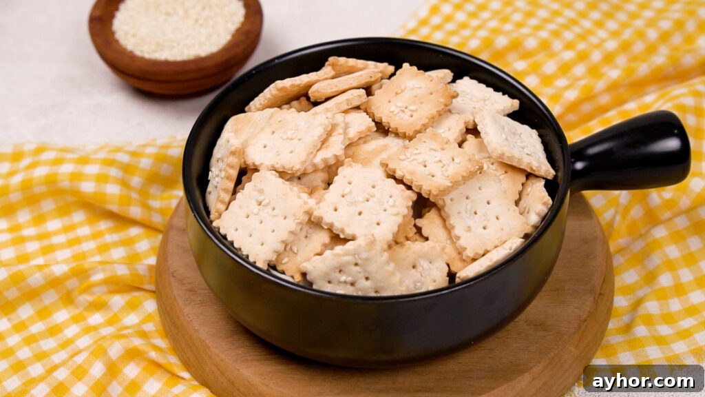 A black bowl filled with golden-brown, square gluten-free crackers topped with sesame seeds, sitting on a wooden board with a yellow gingham cloth.