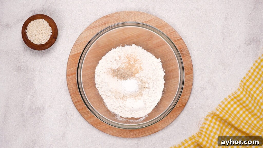 A glass mixing bowl filled with gluten-free flour, sesame seeds, garlic powder, baking powder, and other dry ingredients, ready to be stirred.