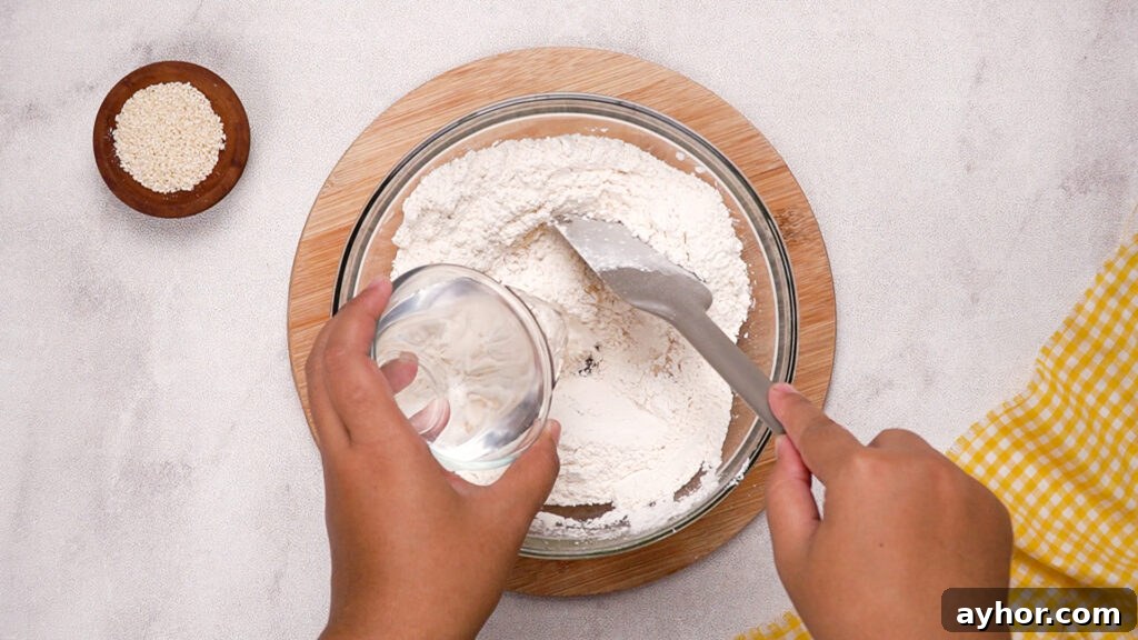 A hand pouring water from a glass cup into the flour mixture while another hand holds a spatula, starting to form the gluten-free cracker dough.