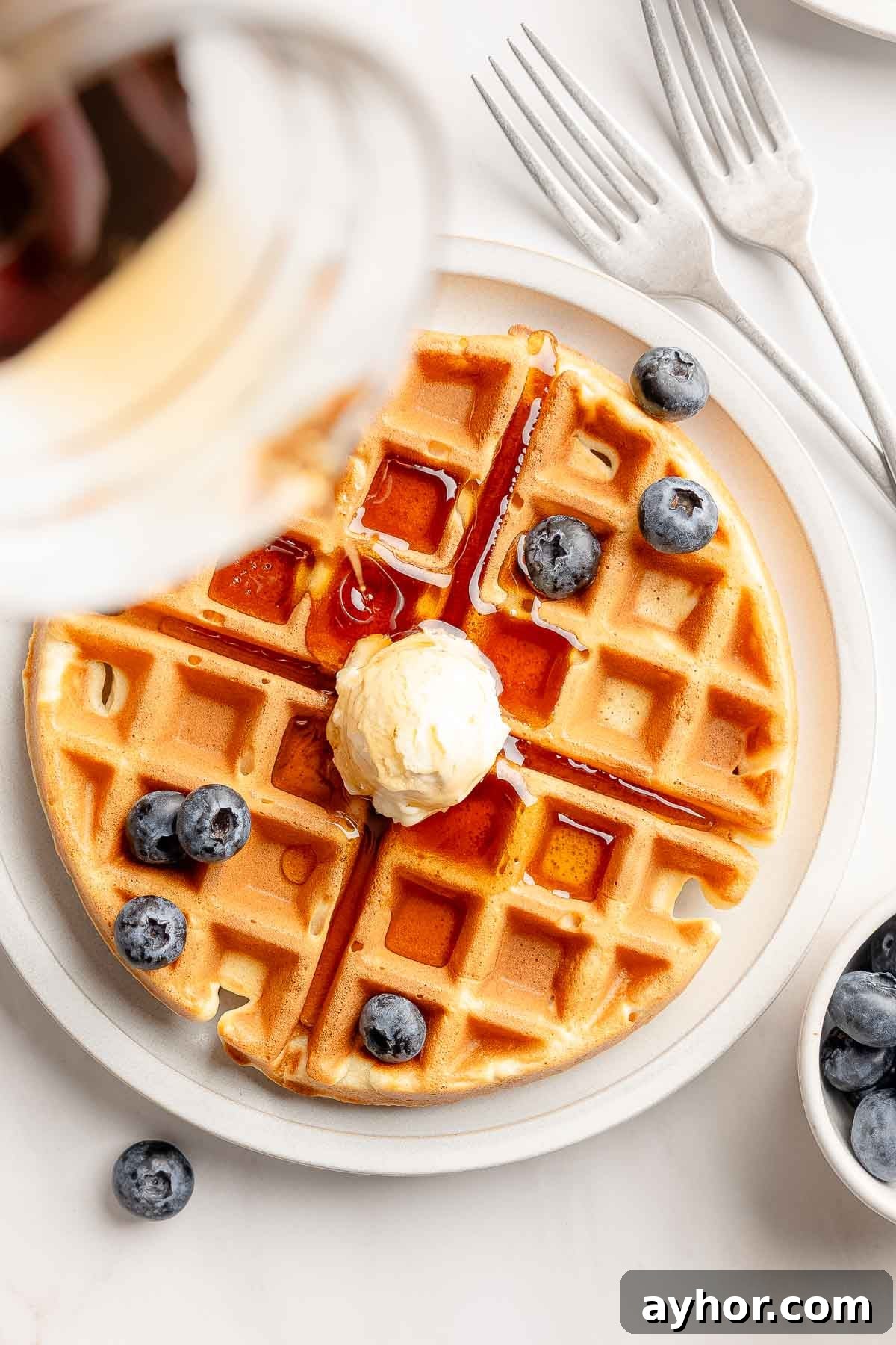 Overhead view of gluten-free waffles garnished with butter and blueberries, is drizzled with syrup.