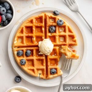 Overhead view of a gluten-free Belgian waffle on a white plate topped with whipped butter, maple syrup, and fresh blueberries, with a bite taken out on a fork.